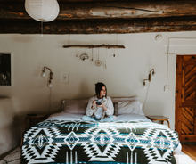 Load image into Gallery viewer, Person sitting on a bed with a patterned blanket in a rustic room.