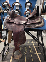 A dark brown tooled leather western saddle displayed on a blue stand in a showroom. The saddle features intricate floral carving and silver-toned metal accents, with matching leather hanging from the bit ring and footrests.