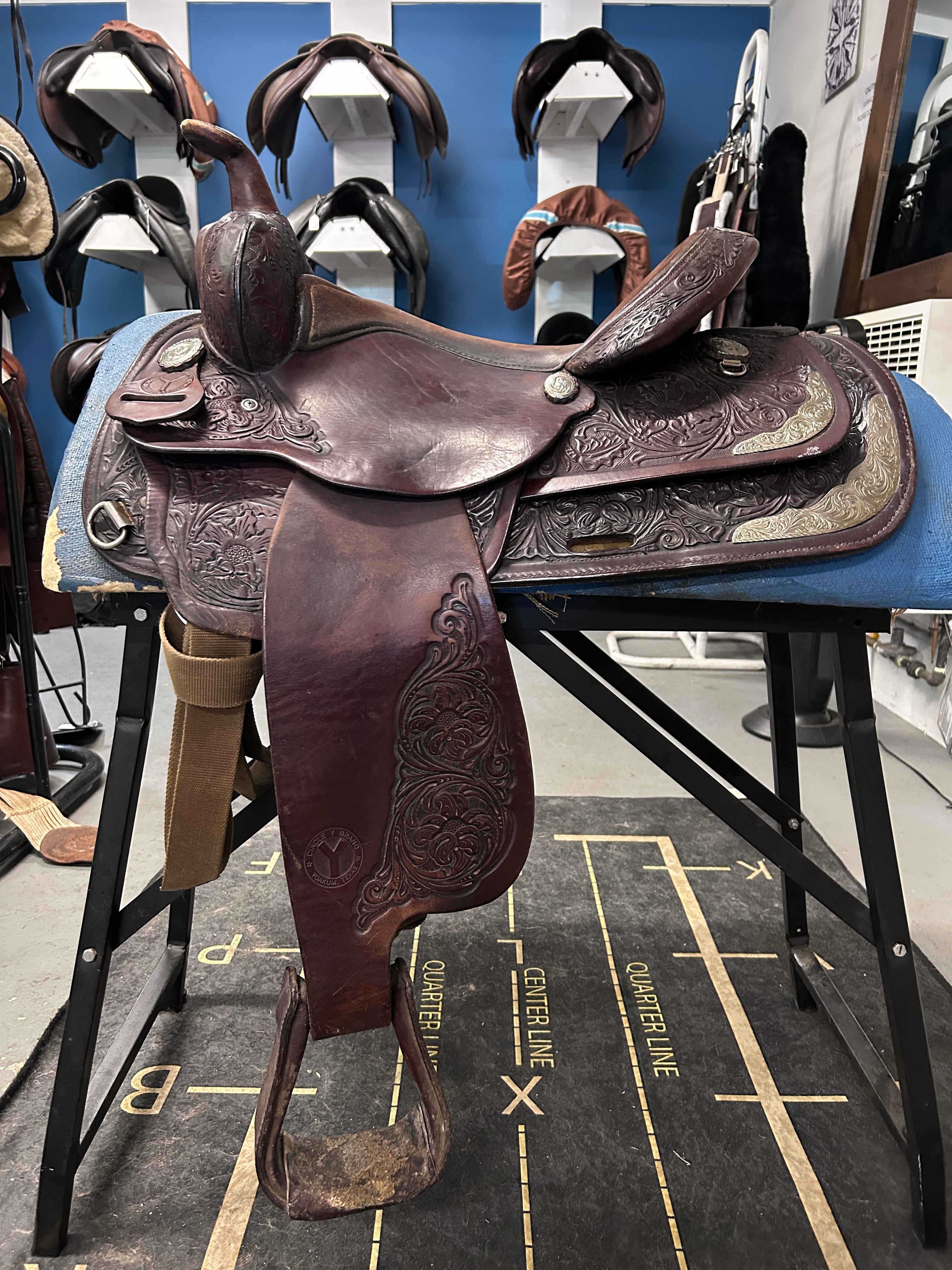 A dark brown tooled leather western saddle displayed on a blue stand in a showroom. The saddle features intricate floral carving and silver-toned metal accents, with matching leather hanging from the bit ring and footrests.