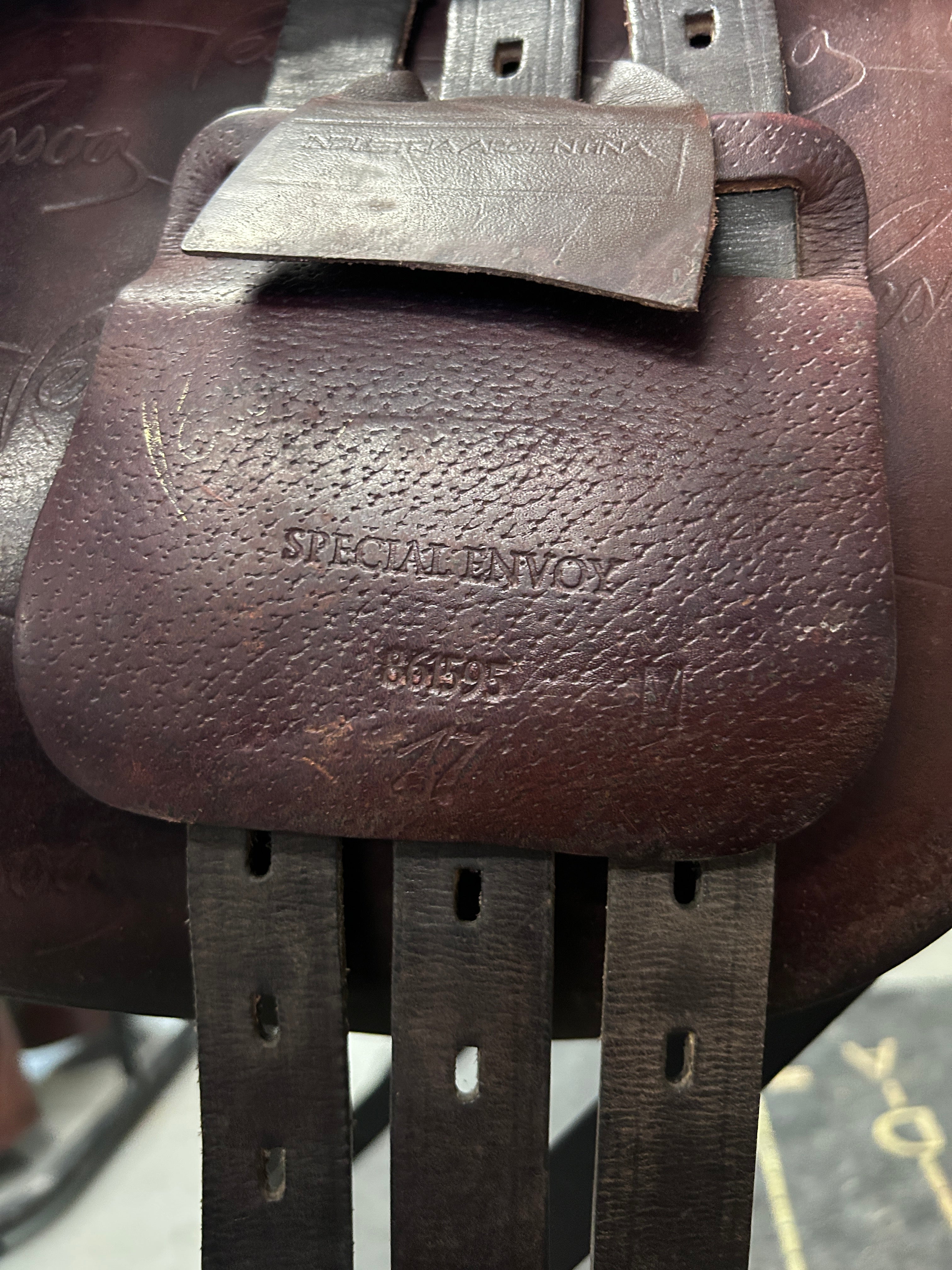 A close-up of a brown leather saddle pad with embossed branding reading 'SPECIAL ENVoy' and 'MEXICO', attached to dark metal saddle straps.