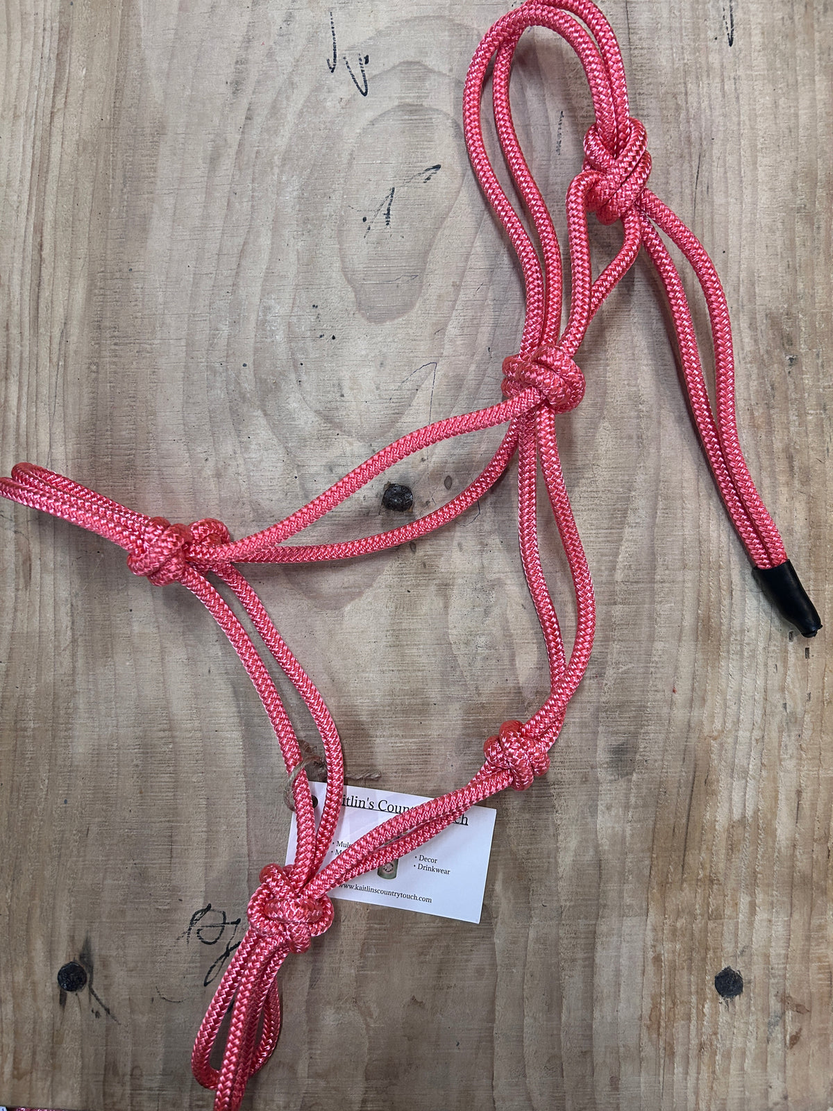 A pink braided rope halter with black nose and mouthpieces, laid flat on a light brown wooden surface with a small white tag attached.