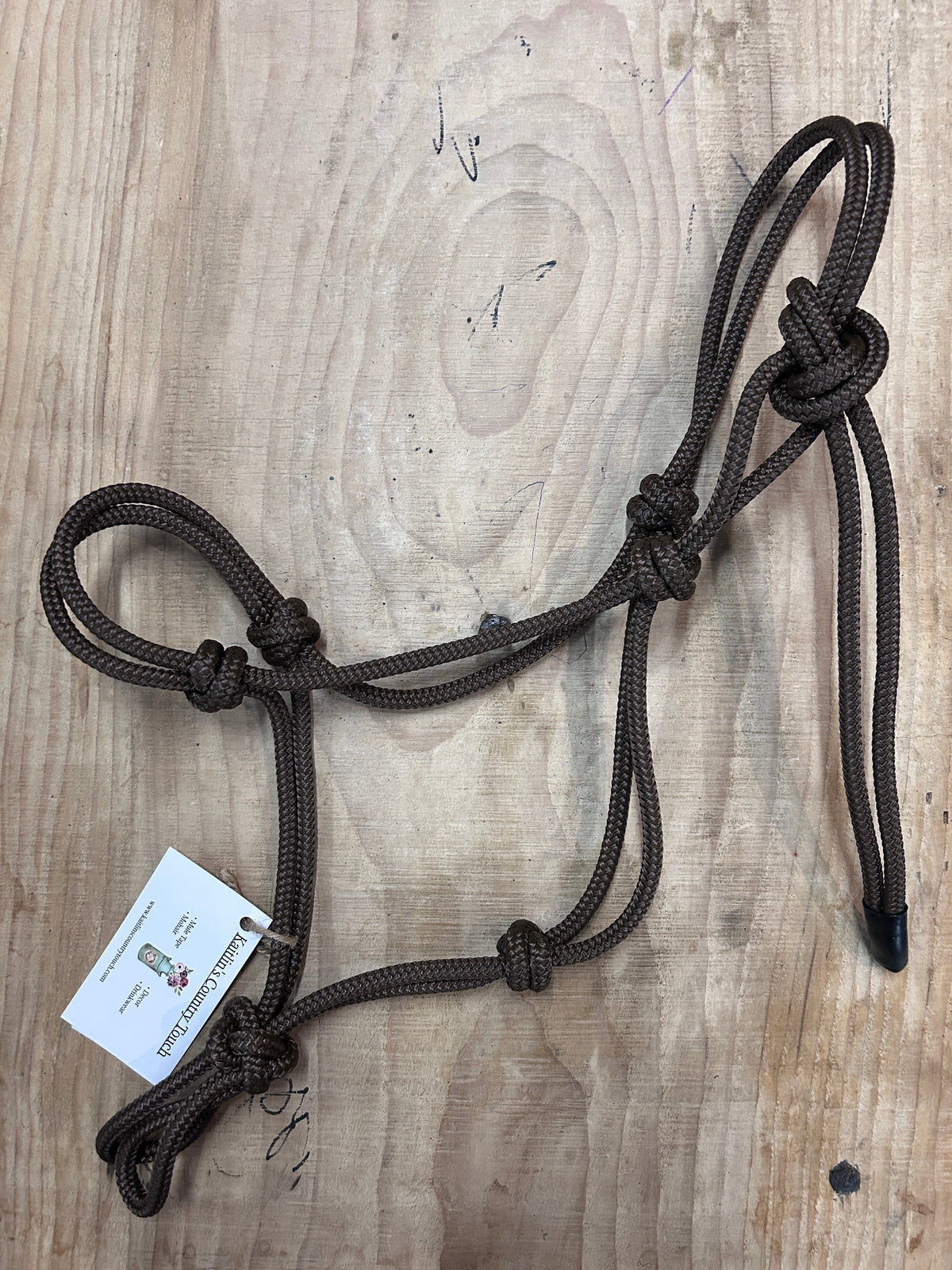A dark brown braided rope halter with a white product tag, laid flat on a light-colored wooden surface.