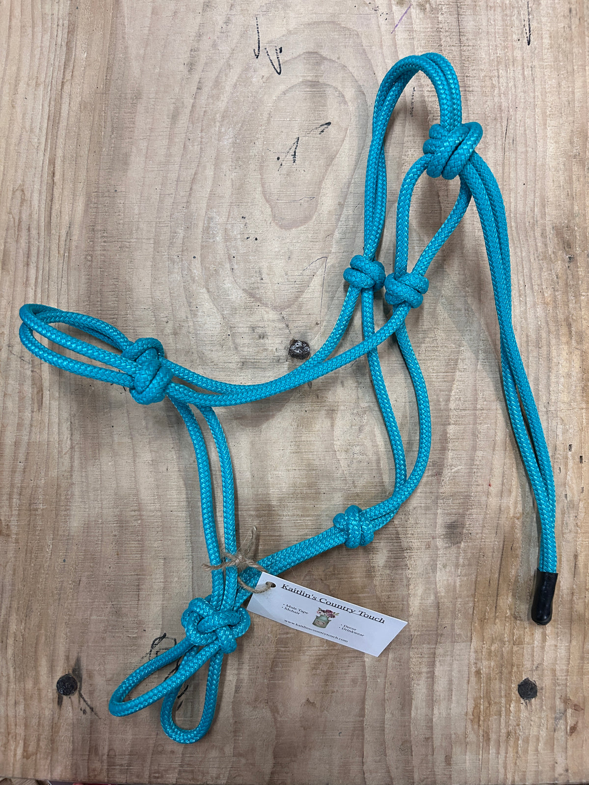 A bright turquoise blue braided rope halter with knots and black noseband tips, laid flat on a light wood grain surface with a small paper tag attached.