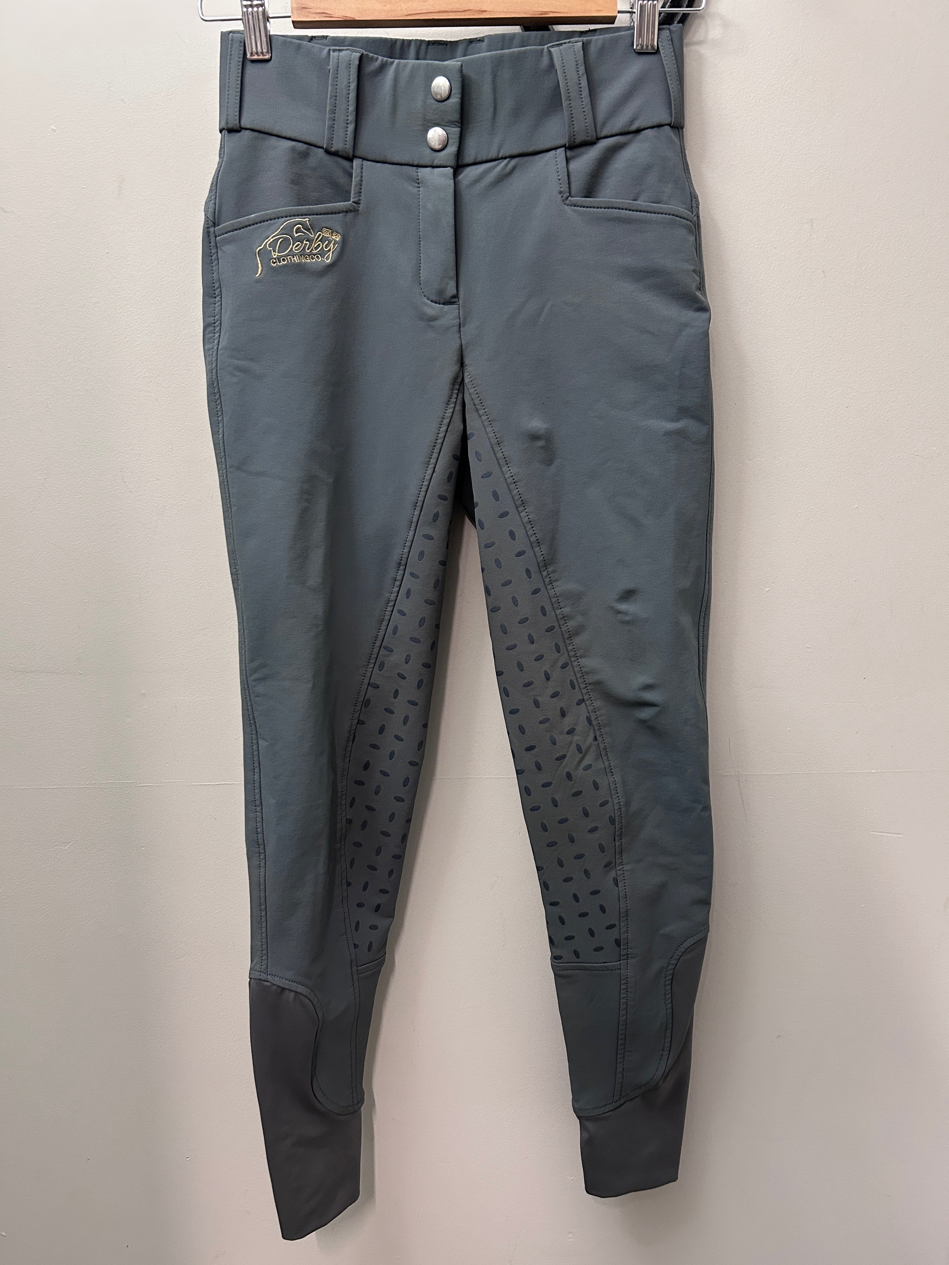 A pair of gray equestrian riding breeches hanging on a wooden hanger against a plain light gray wall. The pants feature a high waist with snap buttons, side pockets, textured grip panels on the lower legs, and a small embroidered logo on the left thigh.