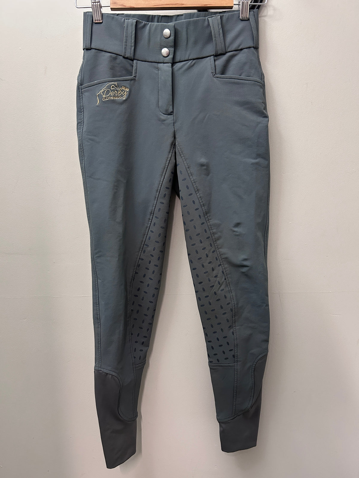 A pair of gray equestrian riding breeches hanging on a wooden hanger against a plain light gray wall. The pants feature a high waist with snap buttons, side pockets, textured grip panels on the lower legs, and a small embroidered logo on the left thigh.