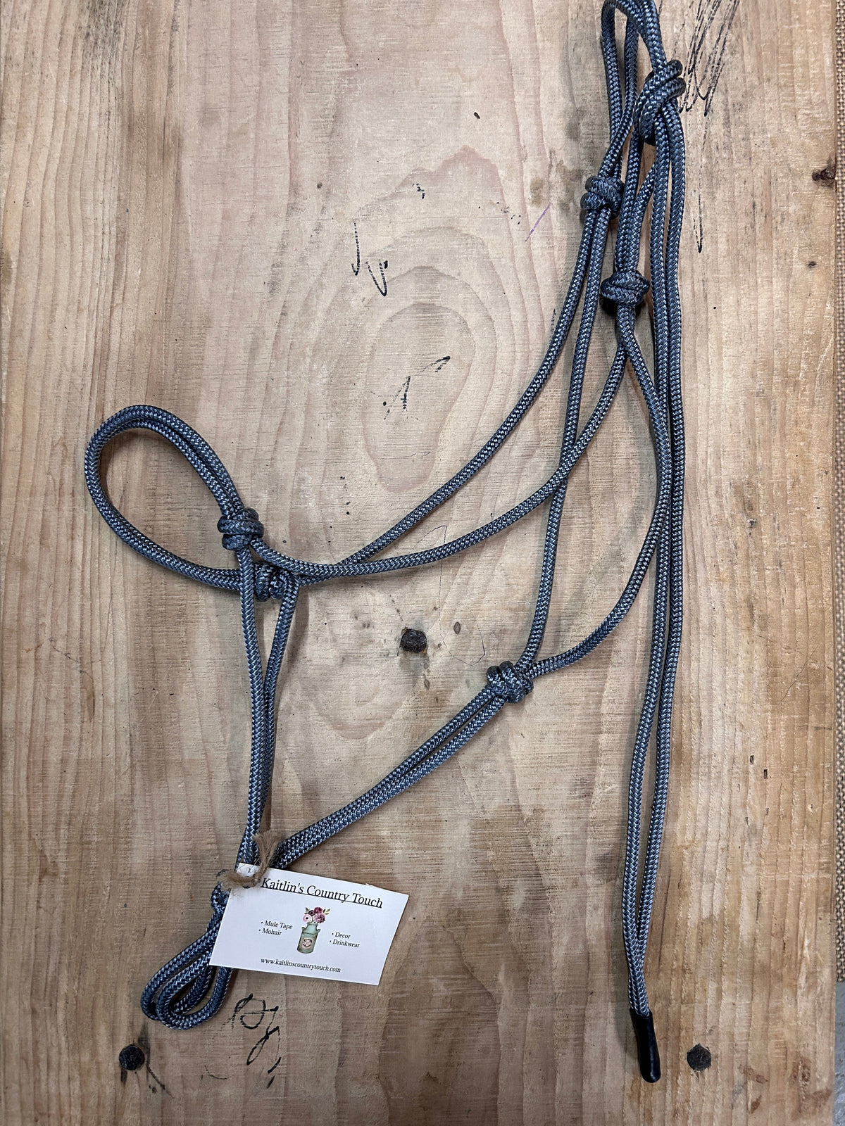 A dark gray braided soft rope halter for horses and a white product tag, laid flat on a light brown wooden surface.