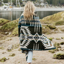 Load image into Gallery viewer, Person wrapped in a patterned blanket standing on a rocky beach with water and greenery in the background
