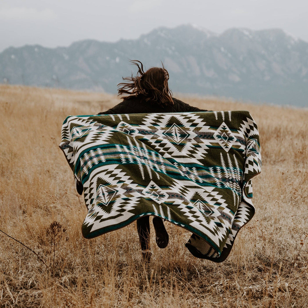 Person with a patterned blanket draped over them in a field with mountains in the background
