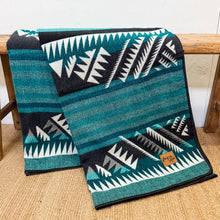 Load image into Gallery viewer, Teal and black patterned blanket with white geometric designs on a wooden surface.
