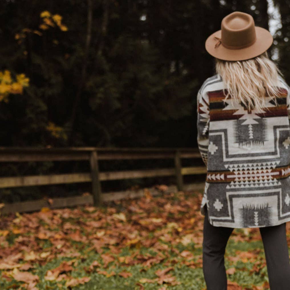 Person wearing a patterned coat and hat standing in an autumn setting with fallen leaves and trees.