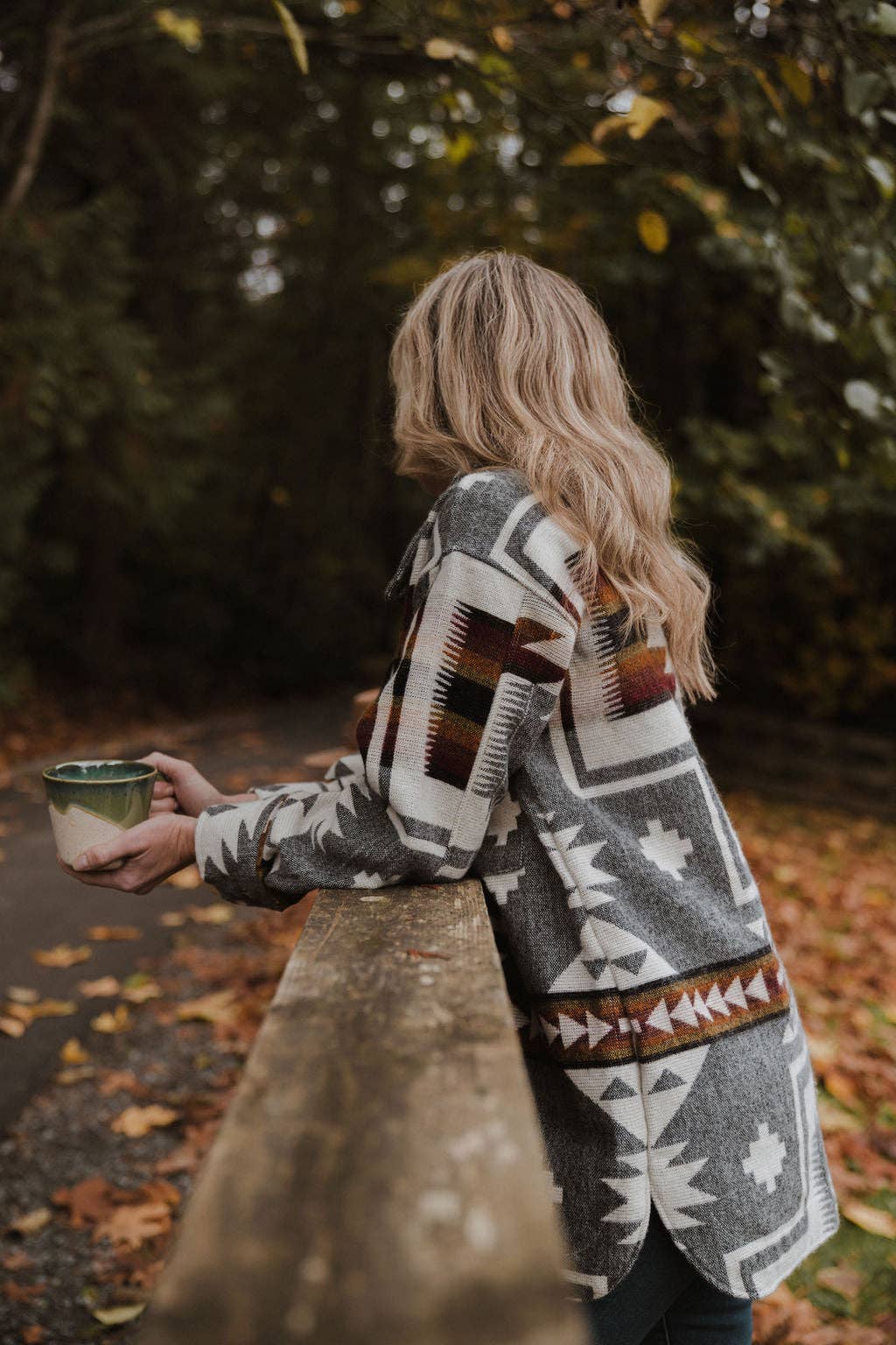 Person wearing a patterned coat holding a mug outdoors with trees in the background